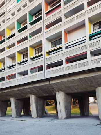A section of a modernist residential building with a grid-like facade. The structure features unevenly sized rectangular windows with different colored panels, including green, yellow, red, and blue. The building is elevated on large concrete pillars, creating an open space underneath.