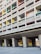 A section of a modernist residential building with a grid-like facade. The structure features unevenly sized rectangular windows with different colored panels, including green, yellow, red, and blue. The building is elevated on large concrete pillars, creating an open space underneath.