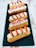 A row of four éclairs is arranged on a dark slate serving tray. Each pastry is garnished with cream, fresh strawberry slices, and pink crumbled toppings. The éclairs have a golden-brown hue and are set against a rustic white wooden background.