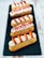 A row of four éclairs is arranged on a dark slate serving tray. Each pastry is garnished with cream, fresh strawberry slices, and pink crumbled toppings. The éclairs have a golden-brown hue and are set against a rustic white wooden background.