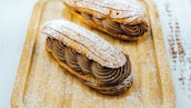 Two éclairs decorated with powdered sugar are placed on a wooden tray. The éclairs are filled with a rich, dark chocolate cream, piped with artistic swirls. The surface of the éclairs and the tray are dusted with powdered sugar, adding a delicate touch to the presentation.