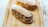 Two éclairs decorated with powdered sugar are placed on a wooden tray. The éclairs are filled with a rich, dark chocolate cream, piped with artistic swirls. The surface of the éclairs and the tray are dusted with powdered sugar, adding a delicate touch to the presentation.
