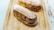 Two éclairs decorated with powdered sugar are placed on a wooden tray. The éclairs are filled with a rich, dark chocolate cream, piped with artistic swirls. The surface of the éclairs and the tray are dusted with powdered sugar, adding a delicate touch to the presentation.