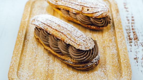 Two &eacute;clairs decorated with powdered sugar are placed on a wooden tray. The &eacute;clairs are filled with a rich, dark chocolate cream, piped with artistic swirls. The surface of the &eacute;clairs and the tray are dusted with powdered sugar, adding a delicate touch to the presentation.