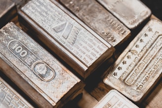 A collection of silver bullion bars, intricately engraved with various inscriptions, stamps, and symbols, stacked closely together. The bars have a reflective surface and display a rich metallic texture.