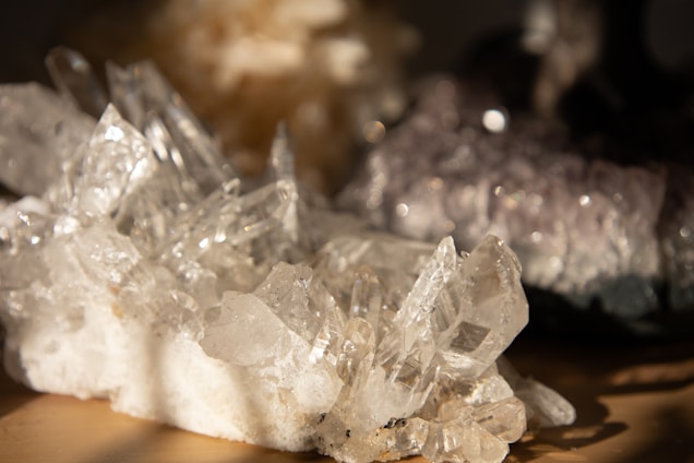 A cluster of clear quartz crystals with sharp, prismatic points, reflecting light with a soft glow. The crystals are resting on a natural base with a texture that contrasts the smooth surfaces of the quartz. In the background, there's a blurred area suggesting other stones or crystals with softer colors.