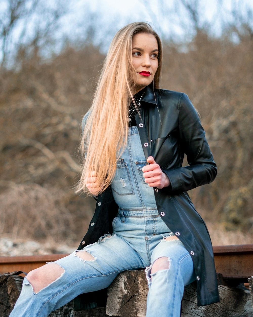 Frau in schwarzer Lederjacke und blauer Jeans sitzt tagsüber auf brauner Holzbank