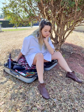 A person sits on an open suitcase, surrounded by brown fallen leaves under a tree in a grassy area. The suitcase contains various clothes and a bottle. The person appears thoughtful or contemplative, wearing a white shirt and brown boots.