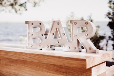 Metallic letters spelling 'BAR' are displayed on a wooden surface, with small round lights embedded in them. The background is blurred but suggests an outdoor setting with trees and water.