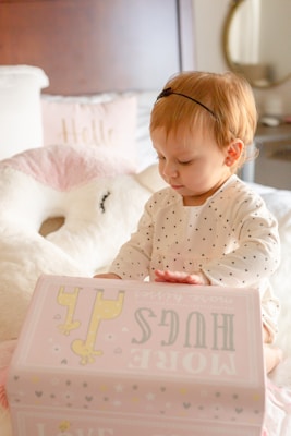 A young child wearing a polka-dotted outfit sits on a bed, intently focusing on a pink box labeled with playful text and illustrations. The background includes soft furnishings like a plush pillow and a bedside mirror.