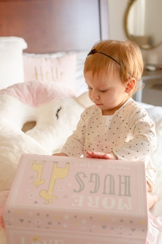 A young child wearing a polka-dotted outfit sits on a bed, intently focusing on a pink box labeled with playful text and illustrations. The background includes soft furnishings like a plush pillow and a bedside mirror.