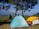 A scenic camping site on a grassy area with two tents set up under tall pine trees. One tent is blue with orange accents, and the other is yellow and green. A person is sitting on a log near the tents, gazing at the expansive landscape of rolling hills and distant trees under a clear blue sky.