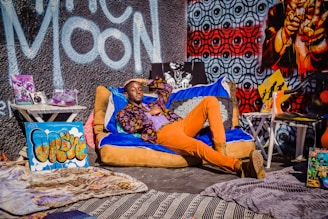 A person is lounging casually on a colorful bean bag chair, surrounded by vibrant street art and eclectic decor. The backdrop features a mix of graffiti art including bold lettering and intricate patterns. Various objects like sneakers, magazines, and art pieces are arranged around, creating a lively and artistic atmosphere.