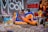 A person is lounging casually on a colorful bean bag chair, surrounded by vibrant street art and eclectic decor. The backdrop features a mix of graffiti art including bold lettering and intricate patterns. Various objects like sneakers, magazines, and art pieces are arranged around, creating a lively and artistic atmosphere.