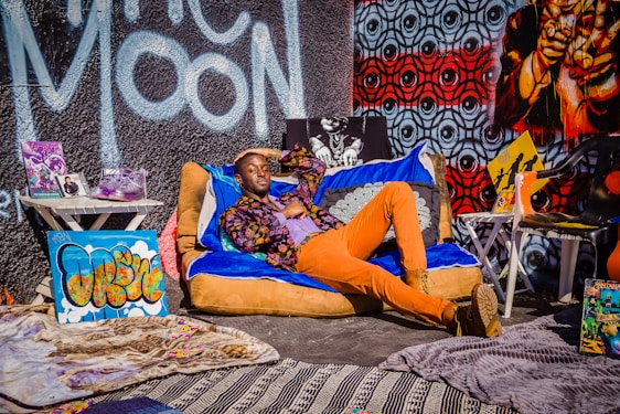 A person is lounging casually on a colorful bean bag chair, surrounded by vibrant street art and eclectic decor. The backdrop features a mix of graffiti art including bold lettering and intricate patterns. Various objects like sneakers, magazines, and art pieces are arranged around, creating a lively and artistic atmosphere.