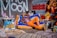 A person is lounging casually on a colorful bean bag chair, surrounded by vibrant street art and eclectic decor. The backdrop features a mix of graffiti art including bold lettering and intricate patterns. Various objects like sneakers, magazines, and art pieces are arranged around, creating a lively and artistic atmosphere.