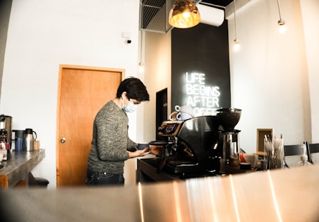 A person wearing a mask is preparing coffee using an espresso machine in a modern cafe. The setting features a neon sign on the wall stating 'Life Begins After Coffee'. The cafe has a minimalist design with warm lighting and a clean counter with utensils and cups.