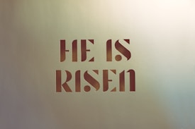 Bold text displays the phrase 'HE IS RISEN' in a modern, stencil-style font against a light textured background.