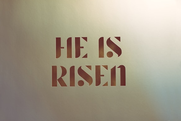 Bold text displays the phrase 'HE IS RISEN' in a modern, stencil-style font against a light textured background.