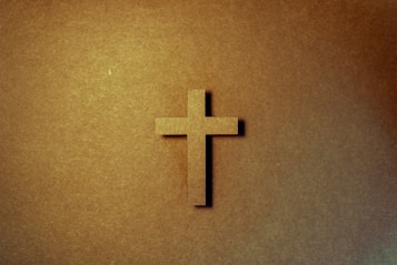 A wooden cross is centered on a textured, warm-toned background, casting a subtle shadow to the right. The lighting creates a soft gradient effect across the image.