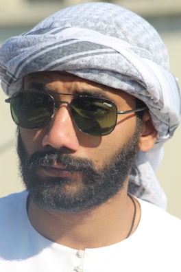 A man with a beard wearing aviator sunglasses and a traditional Middle Eastern headscarf. The scarf is white with subtle patterns. He is dressed in a white garment.