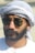 A man with a beard wearing aviator sunglasses and a traditional Middle Eastern headscarf. The scarf is white with subtle patterns. He is dressed in a white garment.