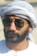 A man with a beard wearing aviator sunglasses and a traditional Middle Eastern headscarf. The scarf is white with subtle patterns. He is dressed in a white garment.