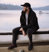 man in black coat sitting on brown wooden bench near body of water during daytime