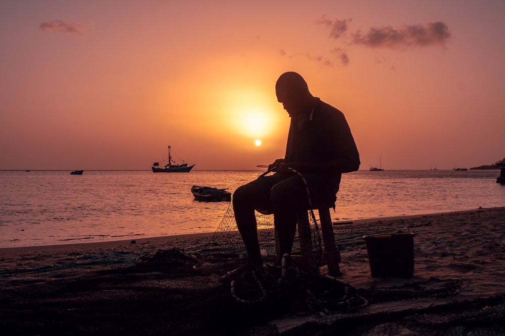 silhouette of man sitting on chair near body of water during sunset