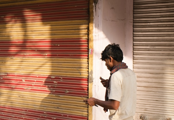 A person stands in front of closed metal shutters, one painted with alternating red and yellow stripes and the other in plain silver. The individual is wearing a light-colored shirt and a scarf around the neck, holding something in their hand. Shadows from nearby objects or people cast intricate patterns on the colorful shutter.