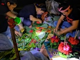 Three individuals are seated on the ground, engaged in crafting decorative arrangements using colorful flowers and green leaves. Their work area is illuminated by a small light source, highlighting the vivid reds, greens, and pinks of the materials. Various crafting supplies, including sticks and plastic bags, are scattered around them.