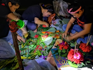 Three individuals are seated on the ground, engaged in crafting decorative arrangements using colorful flowers and green leaves. Their work area is illuminated by a small light source, highlighting the vivid reds, greens, and pinks of the materials. Various crafting supplies, including sticks and plastic bags, are scattered around them.