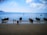 Several traditional longtail boats float on a calm blue sea near the shore. Three people are walking along the sandy beach, and the horizon is lined with distant islands under a partly cloudy sky.
