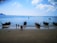 Several traditional longtail boats float on a calm blue sea near the shore. Three people are walking along the sandy beach, and the horizon is lined with distant islands under a partly cloudy sky.