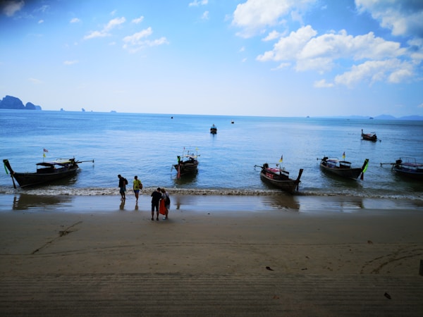Several traditional longtail boats float on a calm blue sea near the shore. Three people are walking along the sandy beach, and the horizon is lined with distant islands under a partly cloudy sky.