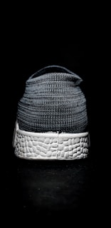 A single shoe is positioned on a dark surface, showcasing its back view. The shoe features a knit-textured upper material in shades of gray, with a prominent white sole characterized by a woven texture.