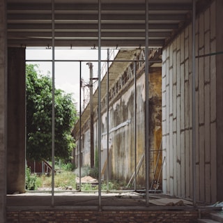An industrial scene with a partially constructed building showing exposed beams and metal framework. The walls are aged with visible rust and decay. A lush green tree contrasts with the industrial elements. There is a mix of rubble and tools scattered on the ground.