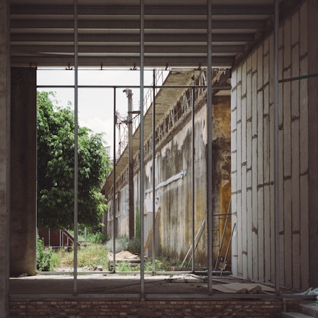 An industrial scene with a partially constructed building showing exposed beams and metal framework. The walls are aged with visible rust and decay. A lush green tree contrasts with the industrial elements. There is a mix of rubble and tools scattered on the ground.