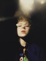 A close-up portrait with a blurred, artistic effect. The person has short curly hair and is wearing glasses. The lighting creates dramatic shadows, and the color palette is soft with dominant dark tones.