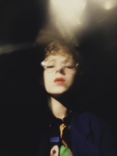 A close-up portrait with a blurred, artistic effect. The person has short curly hair and is wearing glasses. The lighting creates dramatic shadows, and the color palette is soft with dominant dark tones.