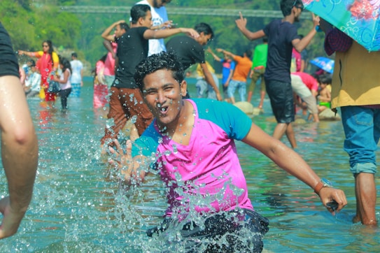 A group of people are enjoying a lively day at a shallow river or beach. The central figure, a young man in a pink and teal shirt, is playfully splashing water towards the camera with a cheerful expression. Around him, other individuals, some with umbrellas and various outfits, are engaging in similar playful activities, creating an atmosphere of fun and leisure. Lush greenery can be seen in the background, and the day appears bright and sunny.