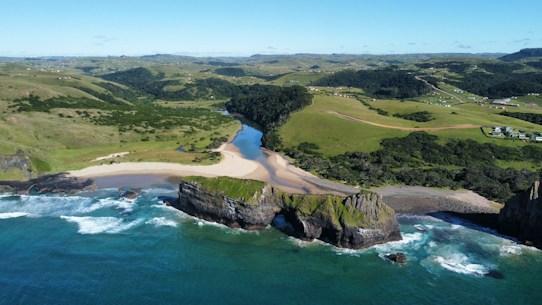 An aerial view of a picturesque coastline featuring a vivid blue ocean meeting a sandy beach, with waves gently crashing on the shore. A large rock formation juts into the sea, covered partly with green vegetation. Behind the beach, a river cuts through lush green hills and distant fields, with a few scattered buildings and roads visible.
