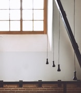 A minimalist interior space featuring a large window with a wooden frame, allowing ample natural light to flood the room. Suspended from the ceiling, several black pendant light fixtures hang at varying lengths. The lower part of the room is accented with a brick wall, adding texture and warmth to the modern design.