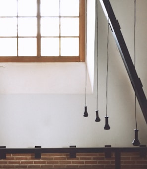A minimalist interior space featuring a large window with a wooden frame, allowing ample natural light to flood the room. Suspended from the ceiling, several black pendant light fixtures hang at varying lengths. The lower part of the room is accented with a brick wall, adding texture and warmth to the modern design.