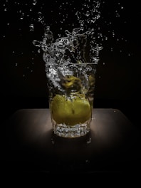 A glass, filled with water, captures the motion of water splashing upwards as a lime is dropped into it. The background is dark, highlighting the splash and the lime.