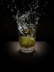 A glass, filled with water, captures the motion of water splashing upwards as a lime is dropped into it. The background is dark, highlighting the splash and the lime.