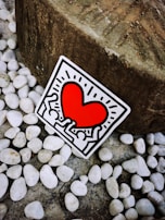 A sticker featuring a vibrant red heart surrounded by abstract human figures outlined in black. The sticker is positioned against a background of smooth white pebbles and a large textured stone.