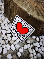 A sticker featuring a vibrant red heart surrounded by abstract human figures outlined in black. The sticker is positioned against a background of smooth white pebbles and a large textured stone.