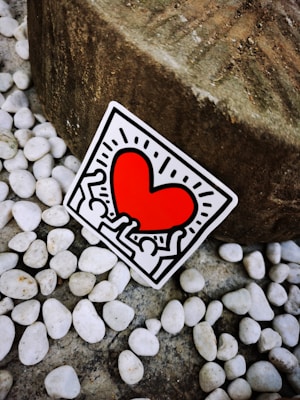 A sticker featuring a vibrant red heart surrounded by abstract human figures outlined in black. The sticker is positioned against a background of smooth white pebbles and a large textured stone.