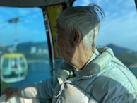 An elderly person with white hair is sitting inside a cable car, wearing a light-colored puffer jacket. The view outside shows a cable car moving along its line, with a scenic backdrop of mountains and a blue sky.